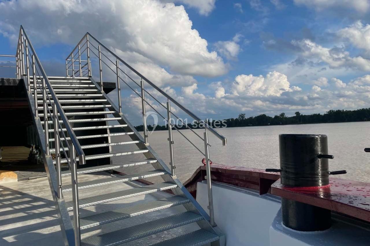 Escalier métallique menant au pont supérieur d'un bateau, avec vue sur un fleuve et un ciel partiellement nuageux.