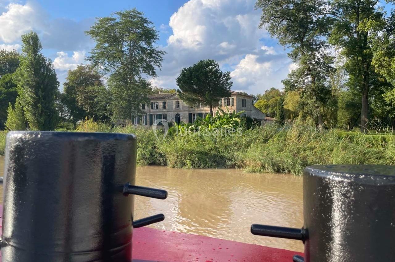 Vue d'une maison en pierre entourée d'arbres, prise depuis un bateau sur une rivière calme.