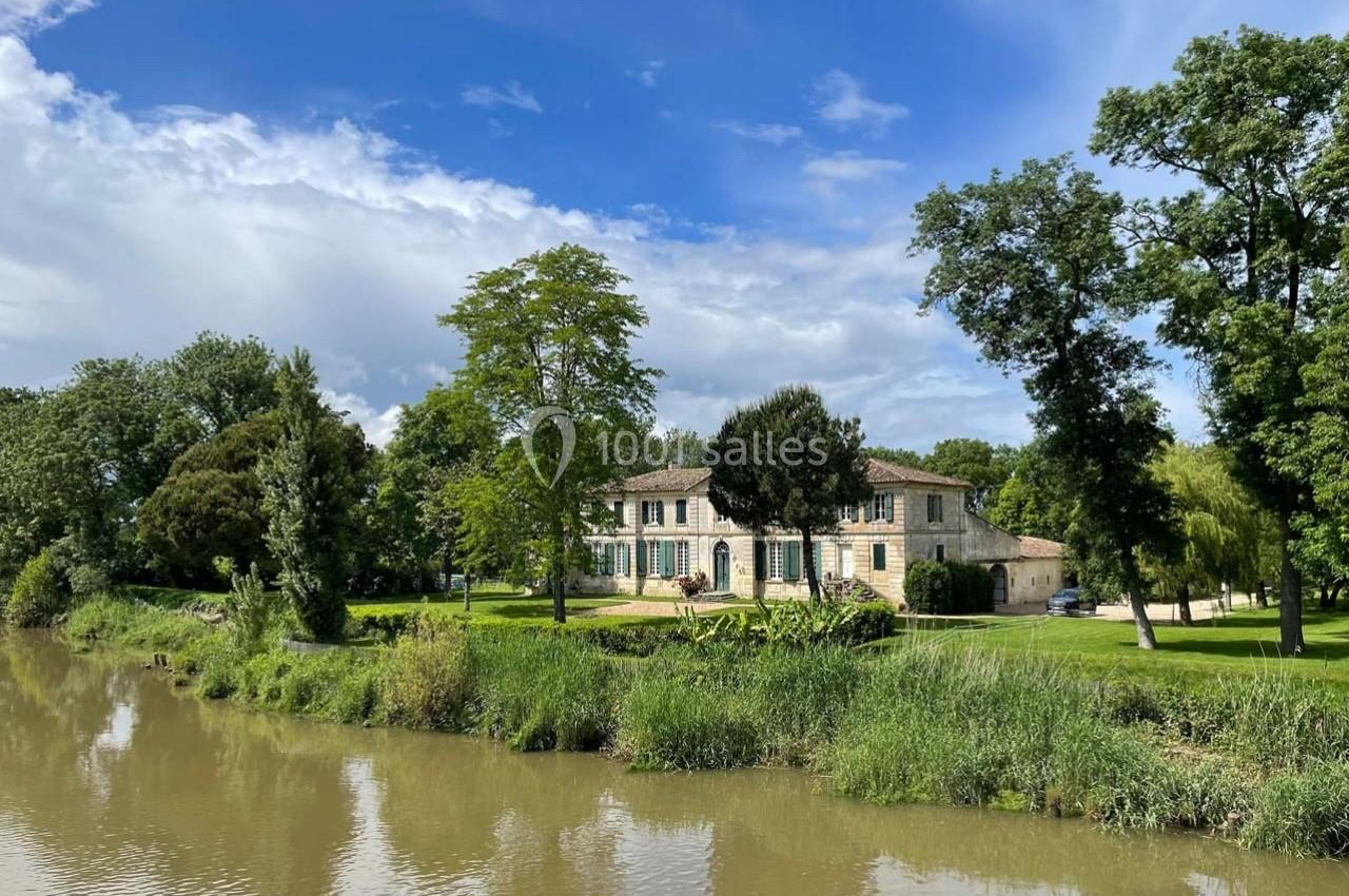 Grande maison en pierre entourée d'arbres et de pelouses, située au bord d'une rivière sous un ciel partiellement nuageux.