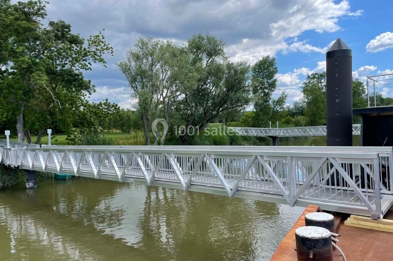 Passerelle métallique traversant une rivière, entourée d'arbres et d'un ciel partiellement nuageux.