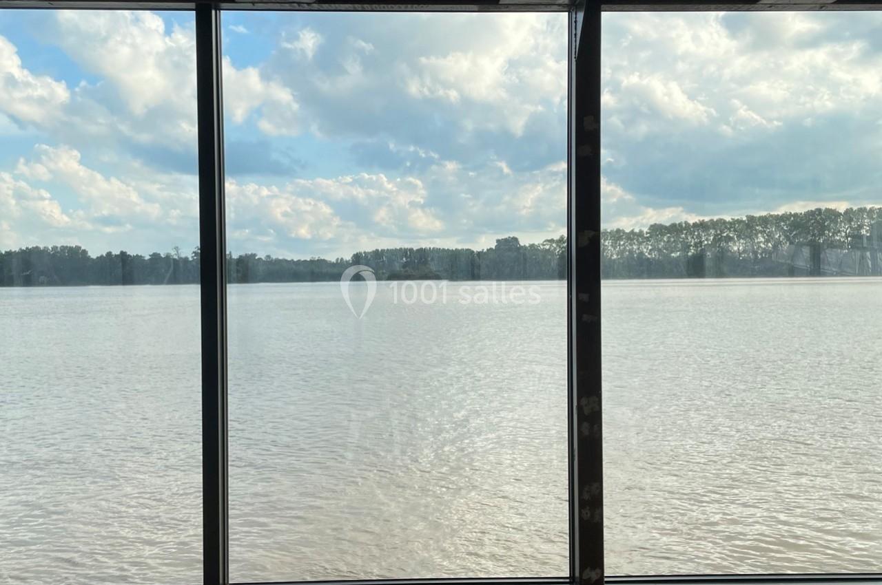Vue d'un large plan d'eau calme bordé d'arbres, observée à travers une baie vitrée par temps nuageux.
