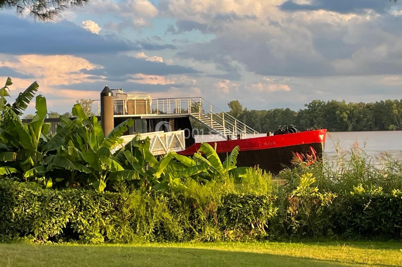 Bateau rouge amarré sur une rivière, entouré de végétation luxuriante sous un ciel partiellement nuageux.