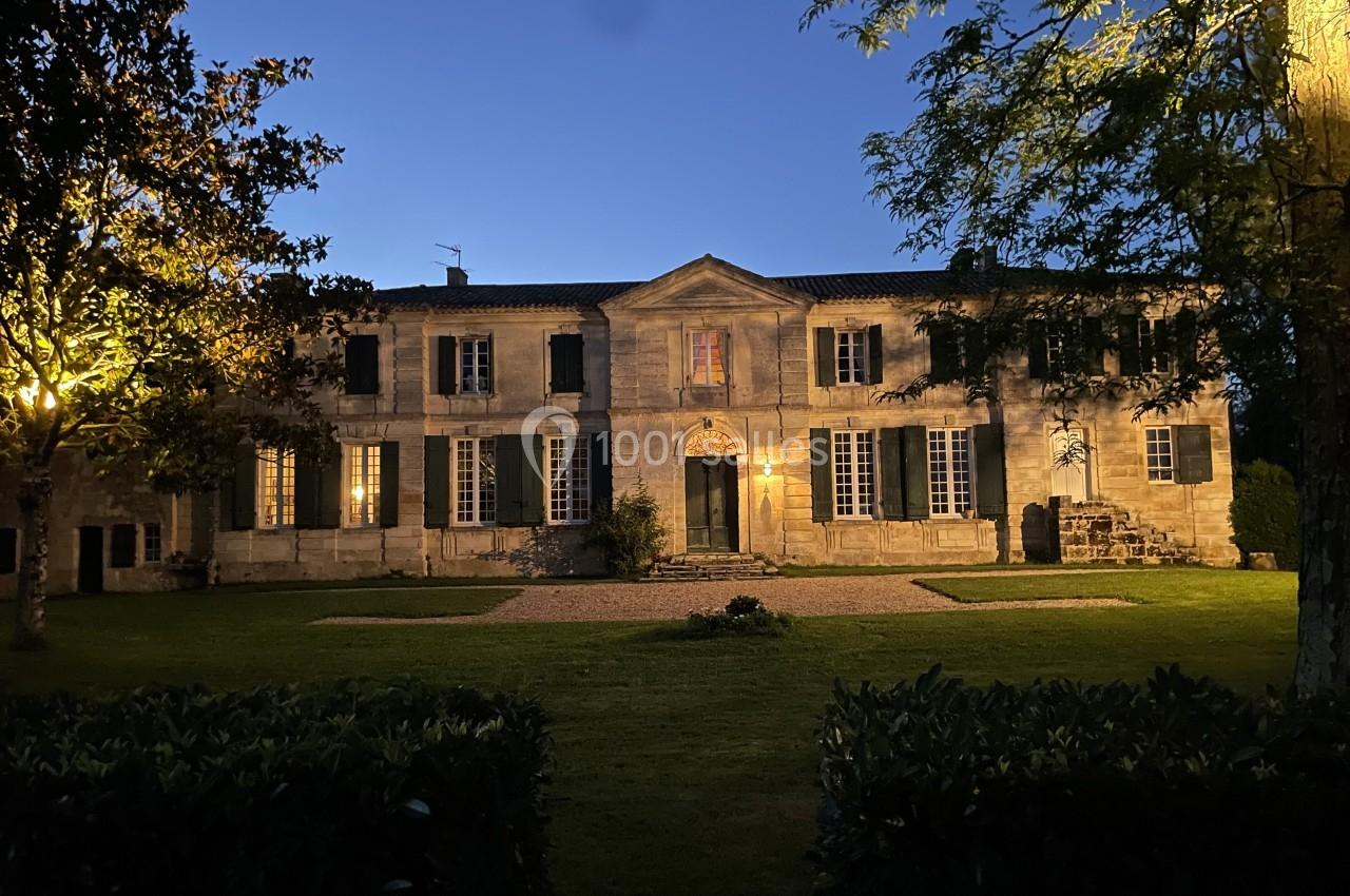 Façade d'une grande maison en pierre éclairée à la tombée de la nuit, entourée d'arbres et d'un jardin.