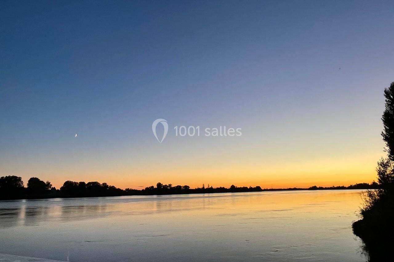 Coucher de soleil sur une rivière calme, avec des silhouettes d'arbres et un croissant de lune visible dans le ciel.