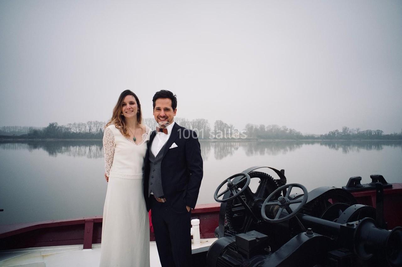 Un couple souriant en tenue élégante pose sur un bateau devant un paysage fluvial brumeux.