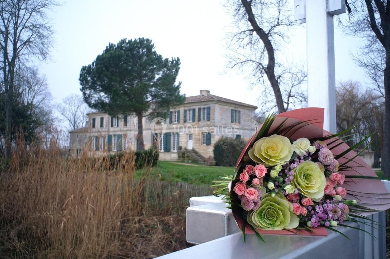 Bouquet de fleurs colorées posé sur une barrière blanche, avec une grande maison en pierre et des arbres en arrière-plan.