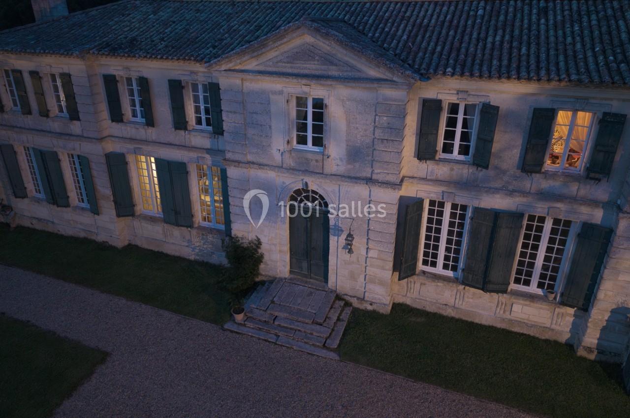 Façade d'une maison en pierre avec volets verts, éclairée de nuit par des lumières intérieures et extérieures.
