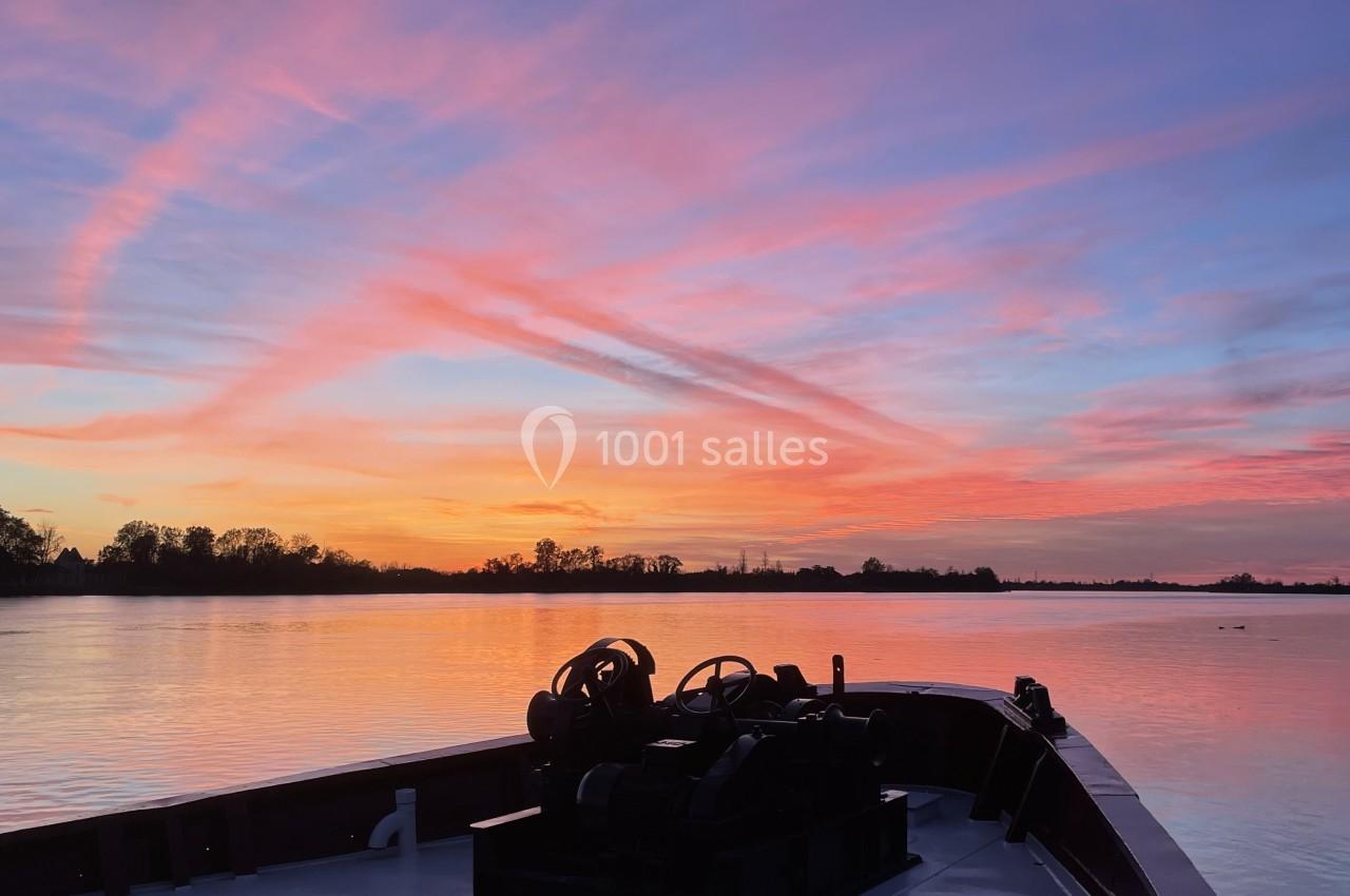 Vue depuis un bateau sur un fleuve au coucher du soleil, avec un ciel coloré de teintes roses et orangées.