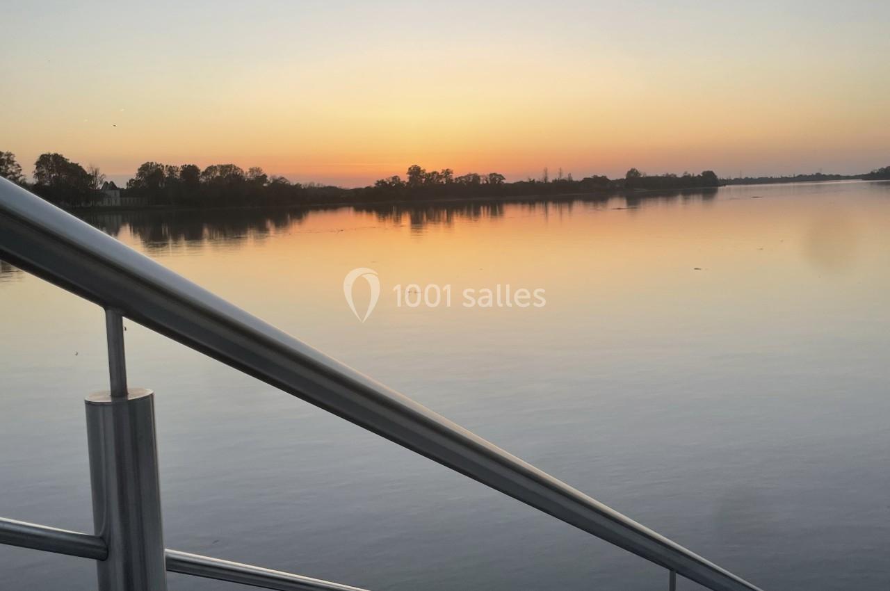 Vue d'un lac calme au coucher du soleil, avec une rambarde métallique au premier plan et des arbres à l'horizon.