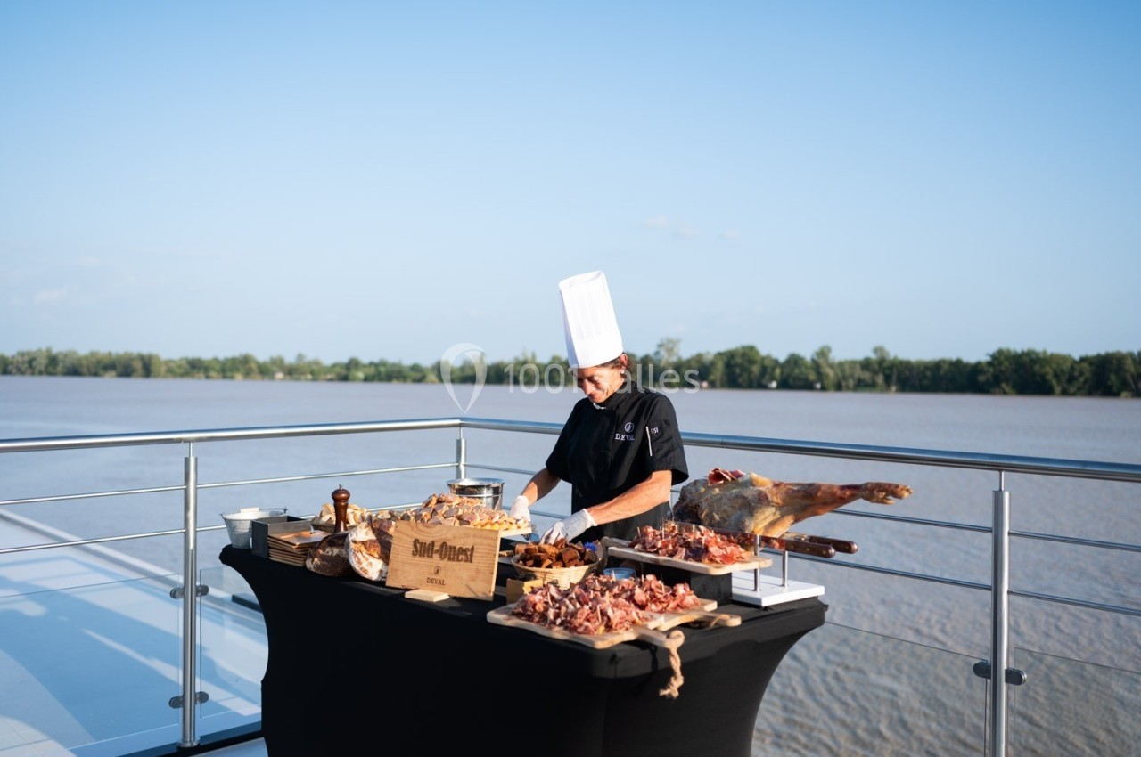 Un chef en tenue professionnelle prépare un buffet de charcuterie sur une terrasse avec vue sur un fleuve.