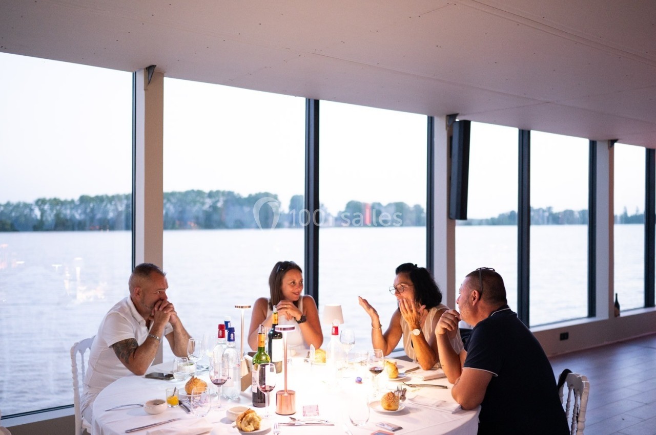 Des personnes discutent autour d'une table dressée dans une salle avec de grandes baies vitrées donnant sur un plan d'eau.