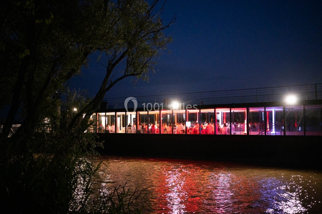 Bâtiment vitré éclairé en rouge et blanc au bord d'une rivière, entouré de végétation, de nuit.