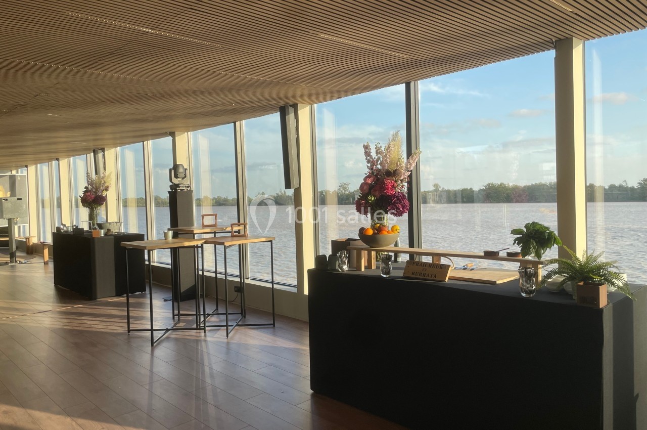Salle lumineuse avec de grandes baies vitrées donnant sur un plan d'eau, tables décorées de fleurs et de fruits.