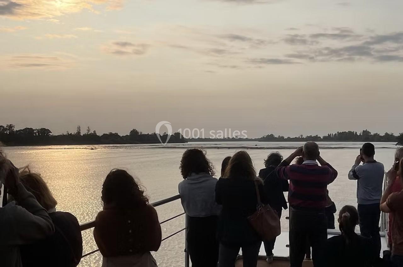 Des personnes observent un paysage fluvial au coucher du soleil depuis le pont d'un bateau.