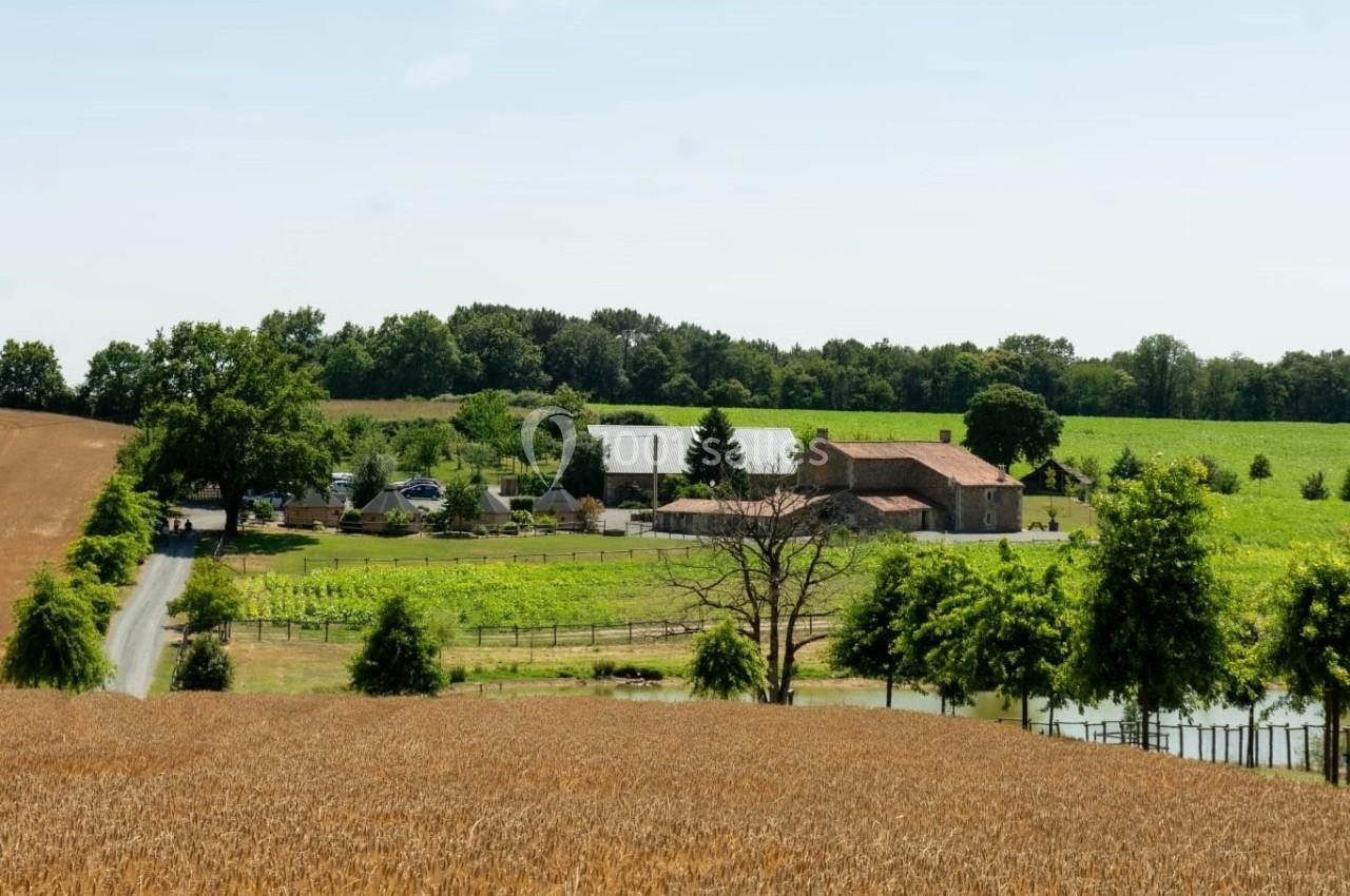 Ferme entourée de champs, d'arbres et de vignes, avec des bâtiments en pierre et un petit étang au premier plan.