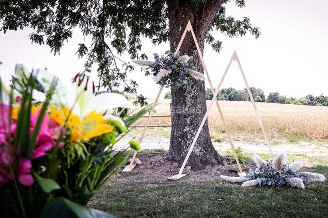 Décor de cérémonie en extérieur avec arche en bois triangulaire, fleurs blanches et champ en arrière-plan.
