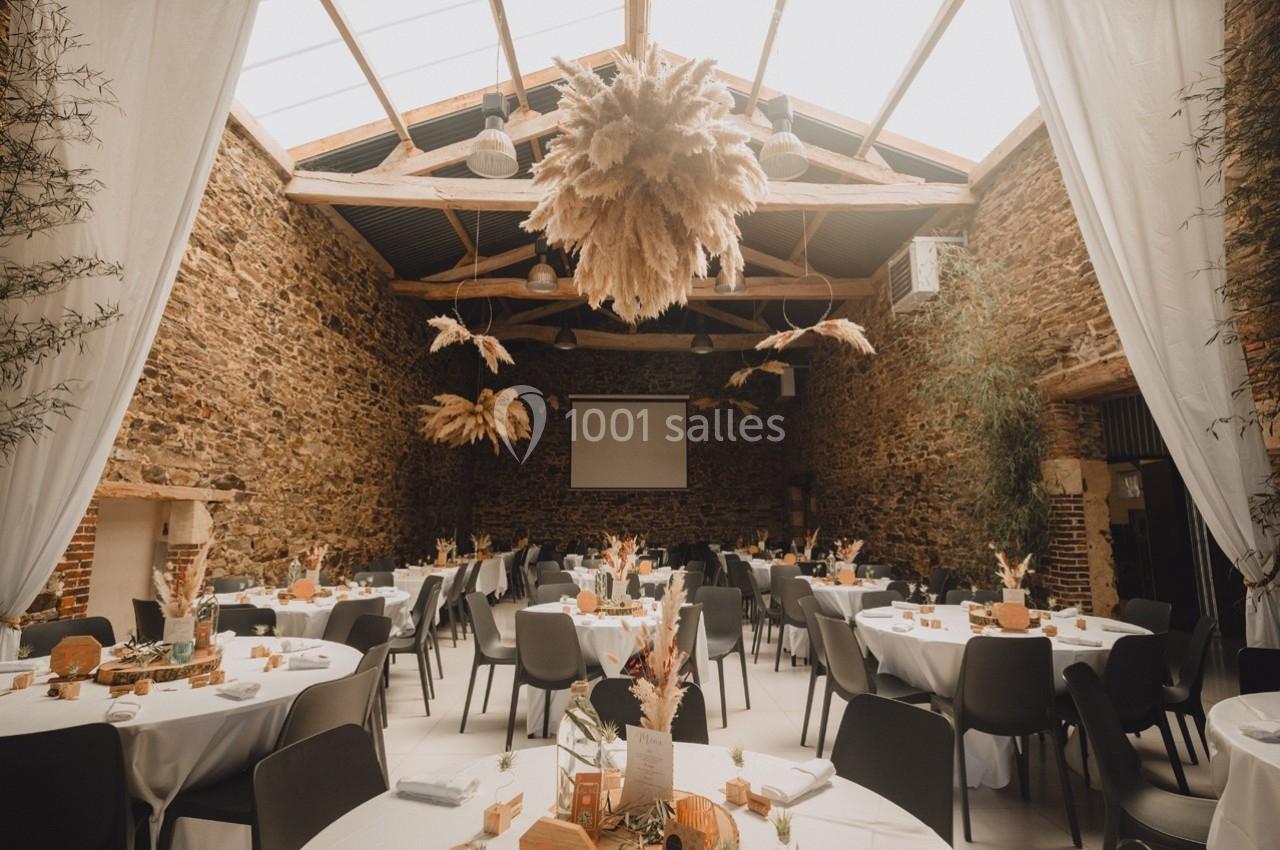 Salle en pierre avec tables rondes dressées, décorations florales et plafond en poutres apparentes.