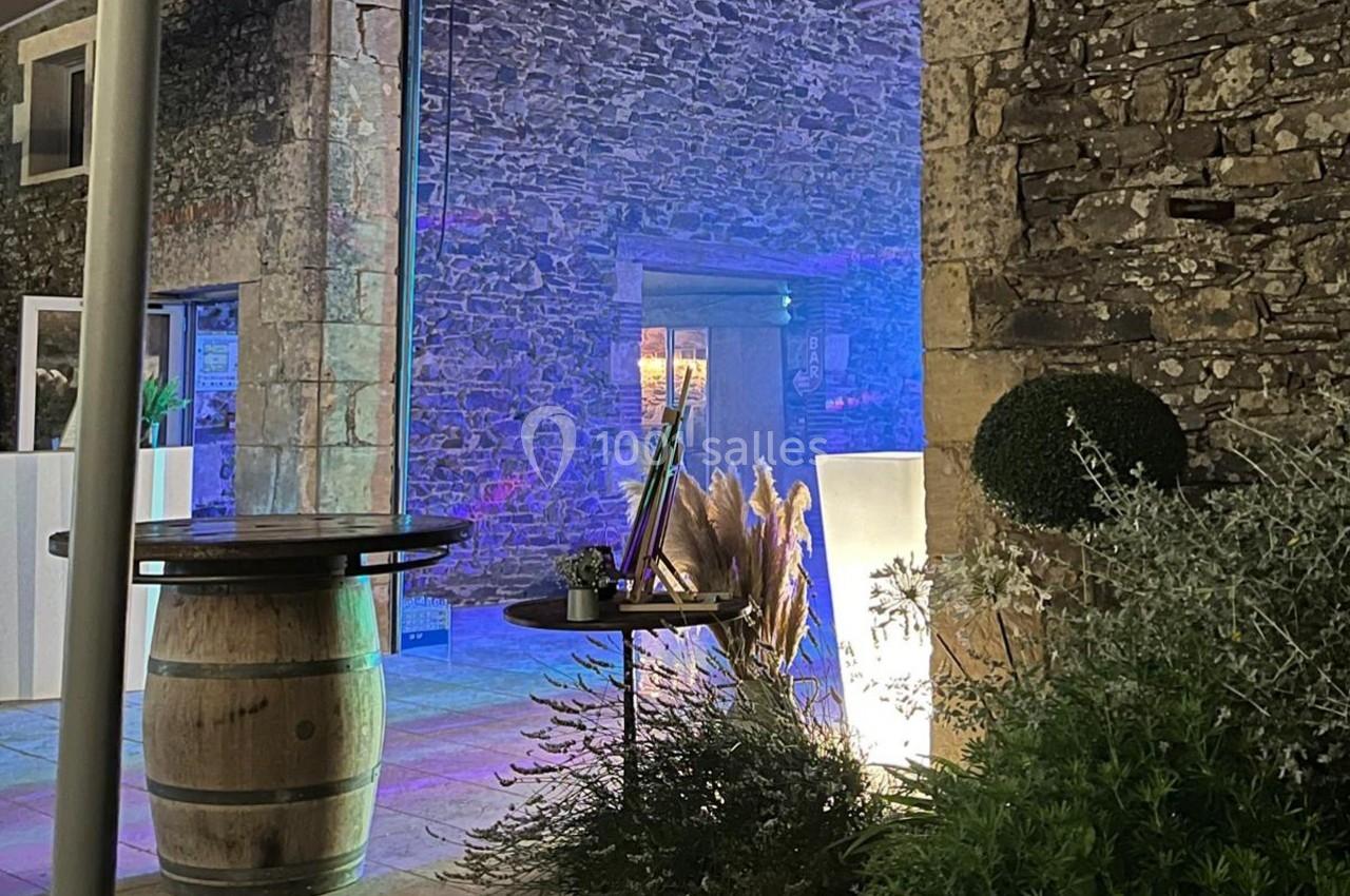 Espace extérieur en soirée avec murs en pierre, végétation, tonneau en bois et éclairage coloré.