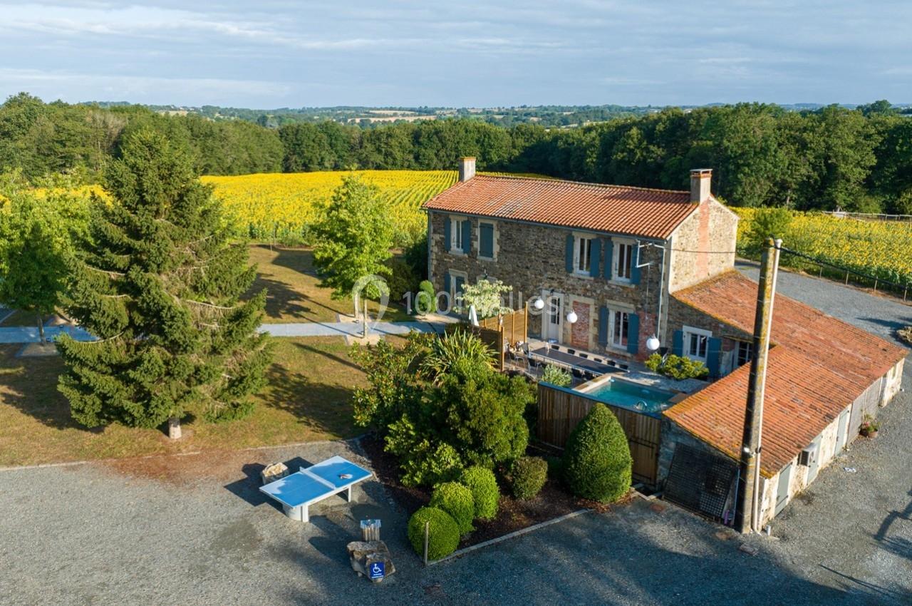 Maison en pierre avec piscine, entourée de champs de tournesols et d'arbres, vue aérienne en journée.