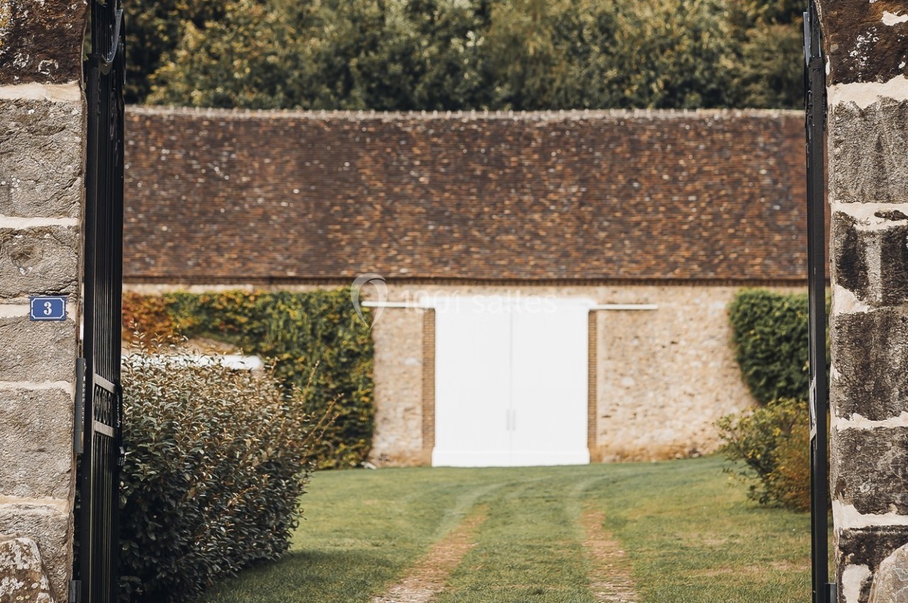 Vue d'une grange en pierre avec une porte blanche, entourée de verdure et encadrée par un portail en pierre.
