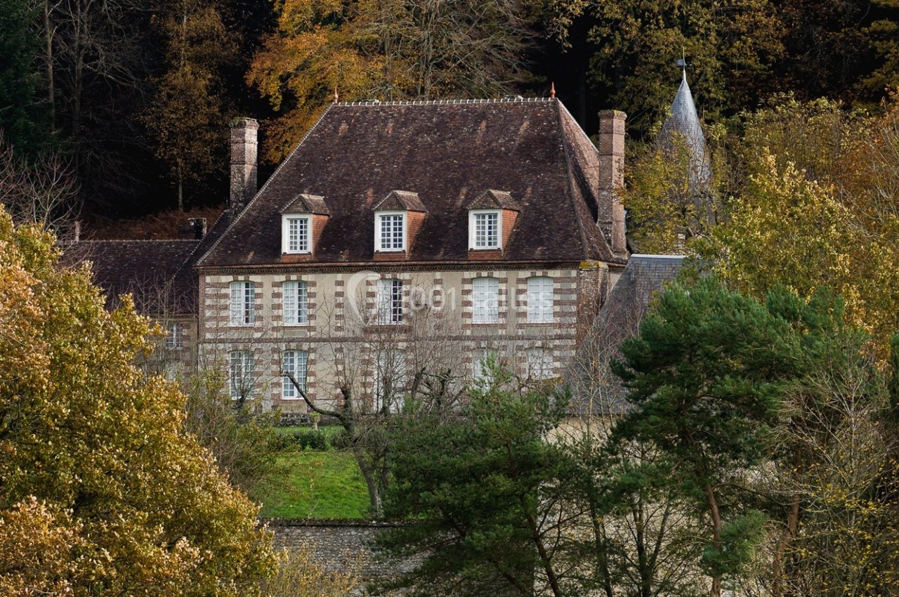 Manoir en pierre entouré d'arbres et de verdure, avec un toit en tuiles et des fenêtres à volets blancs.