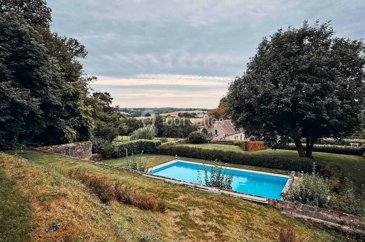 Piscine rectangulaire entourée de verdure, avec vue sur une maison et un paysage vallonné sous un ciel nuageux.