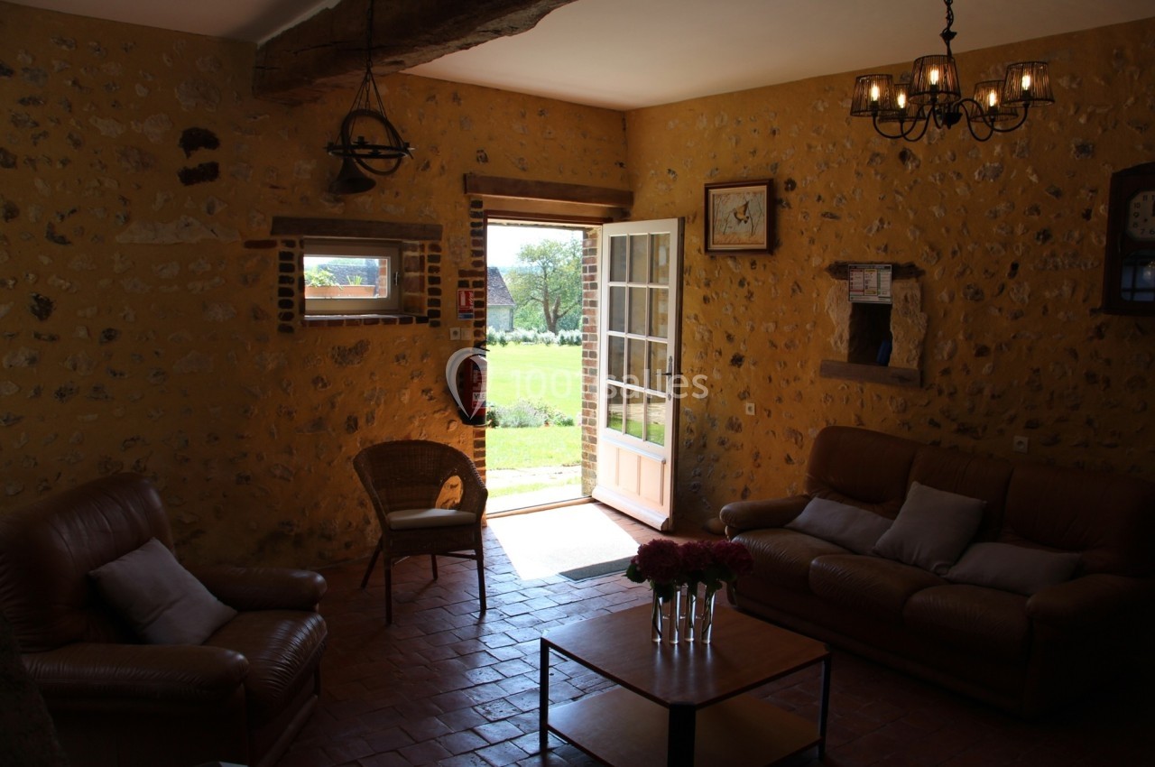 Salon rustique avec murs en pierre, canapés en cuir, table basse et porte ouverte donnant sur un jardin ensoleillé.