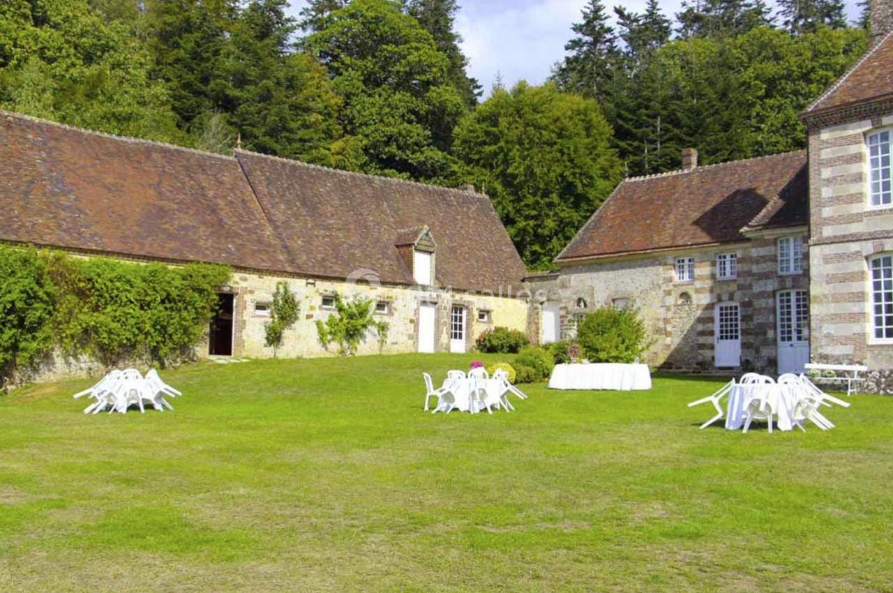Cour verdoyante avec bâtiments en pierre, chaises et tables blanches disposées pour un événement en extérieur.