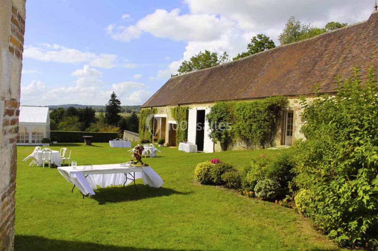 Jardin verdoyant avec tables dressées devant un bâtiment en pierre couvert de lierre, sous un ciel partiellement nuageux.