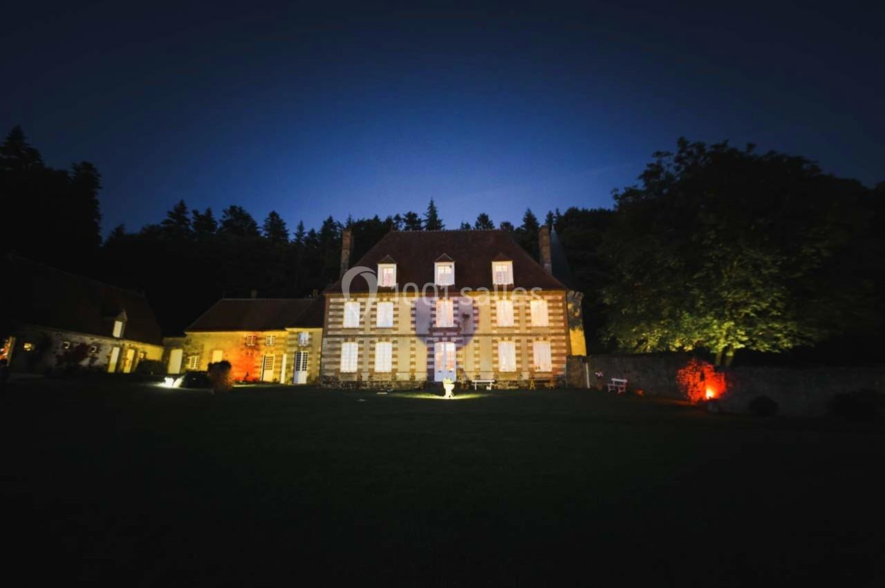 Façade éclairée d'un grand bâtiment ancien entouré d'arbres, photographiée de nuit avec un éclairage coloré.
