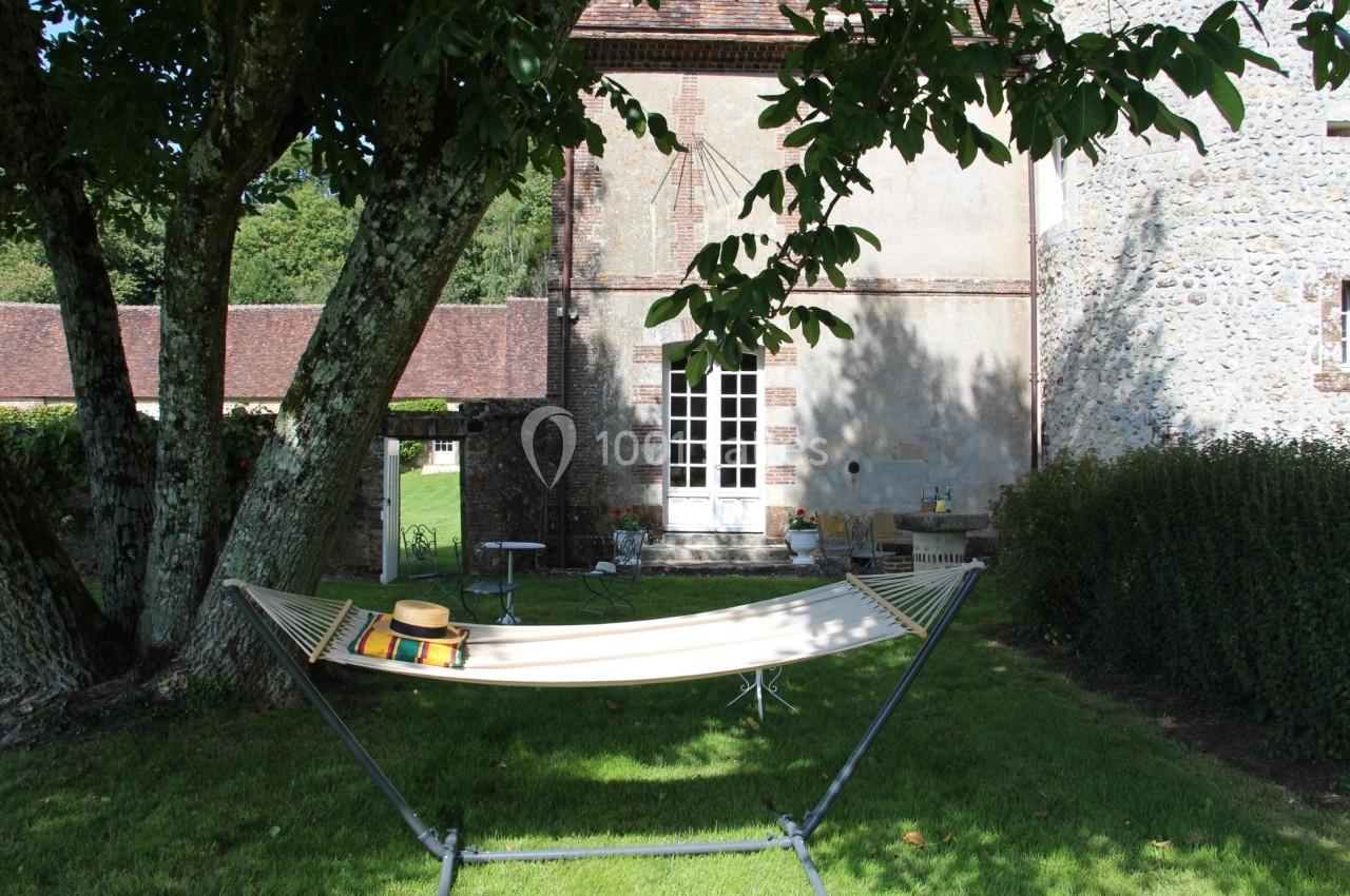 Hamac beige installé sous un arbre dans un jardin verdoyant, près d'une maison en pierre avec des volets blancs.