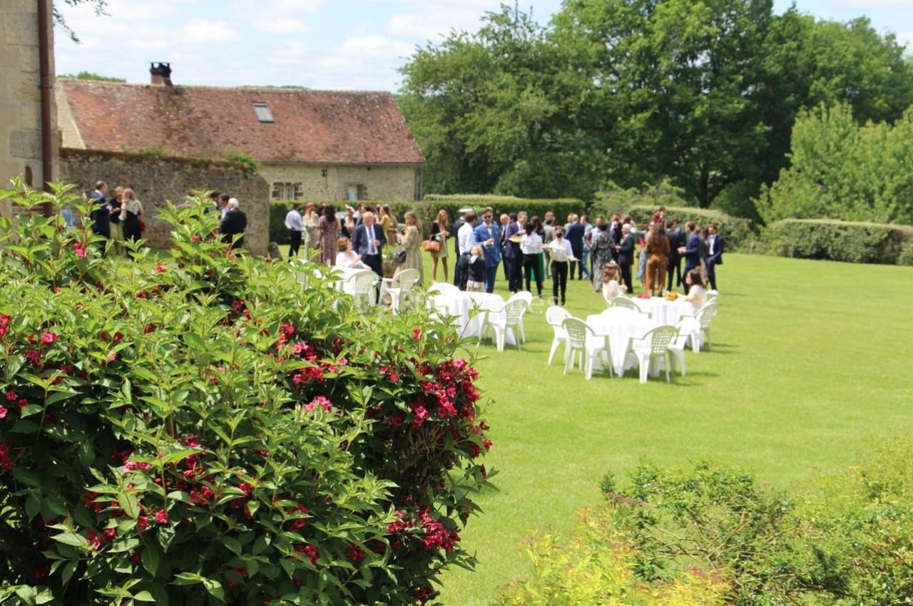 Groupe de personnes rassemblées sur une pelouse avec des chaises blanches, près d'un bâtiment en pierre et d'arbustes…
