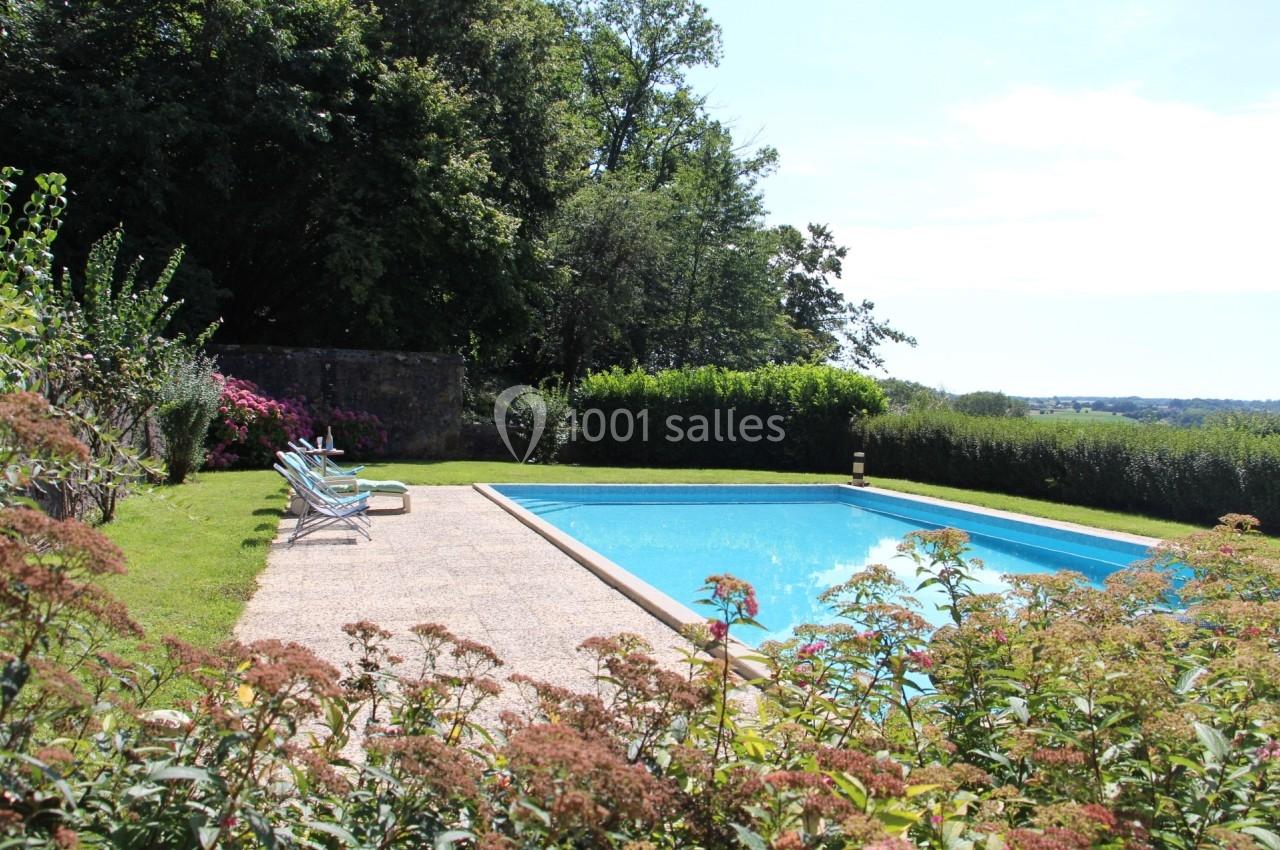 Piscine extérieure entourée de verdure, avec des transats sur une terrasse ensoleillée et un jardin fleuri.