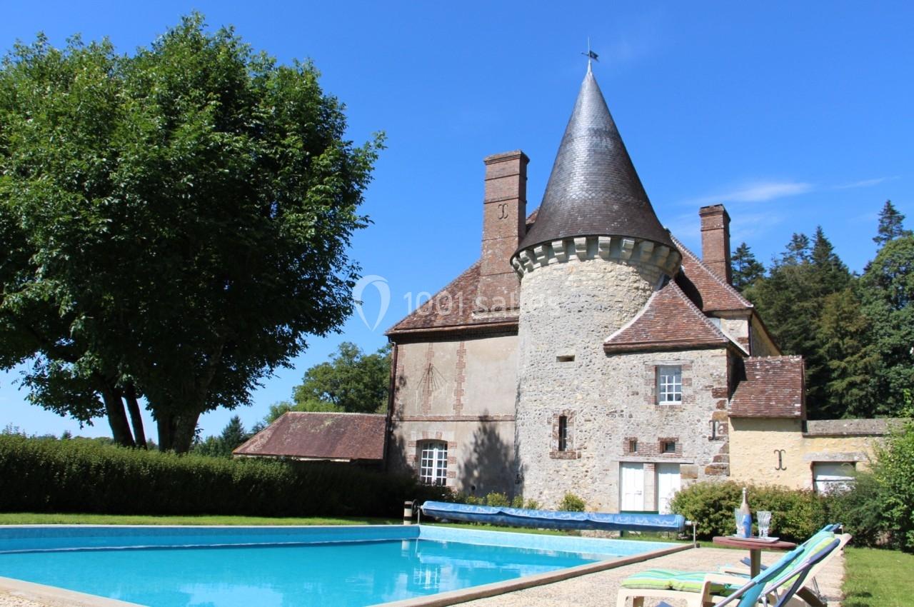 Manoir en pierre avec tour conique, entouré de verdure, avec une piscine au premier plan sous un ciel bleu.