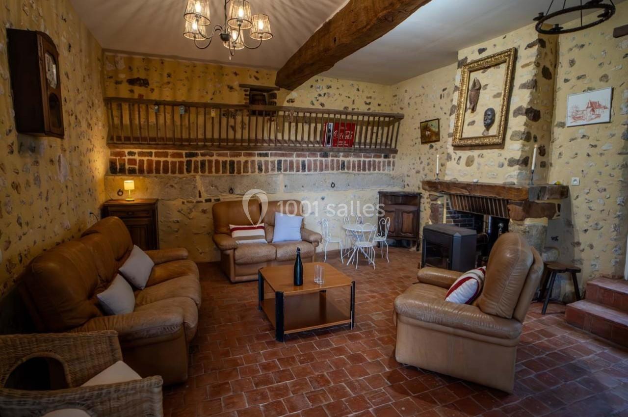 Salon rustique avec murs en pierre, poutres apparentes, mobilier en cuir, cheminée et table en fer forgé.