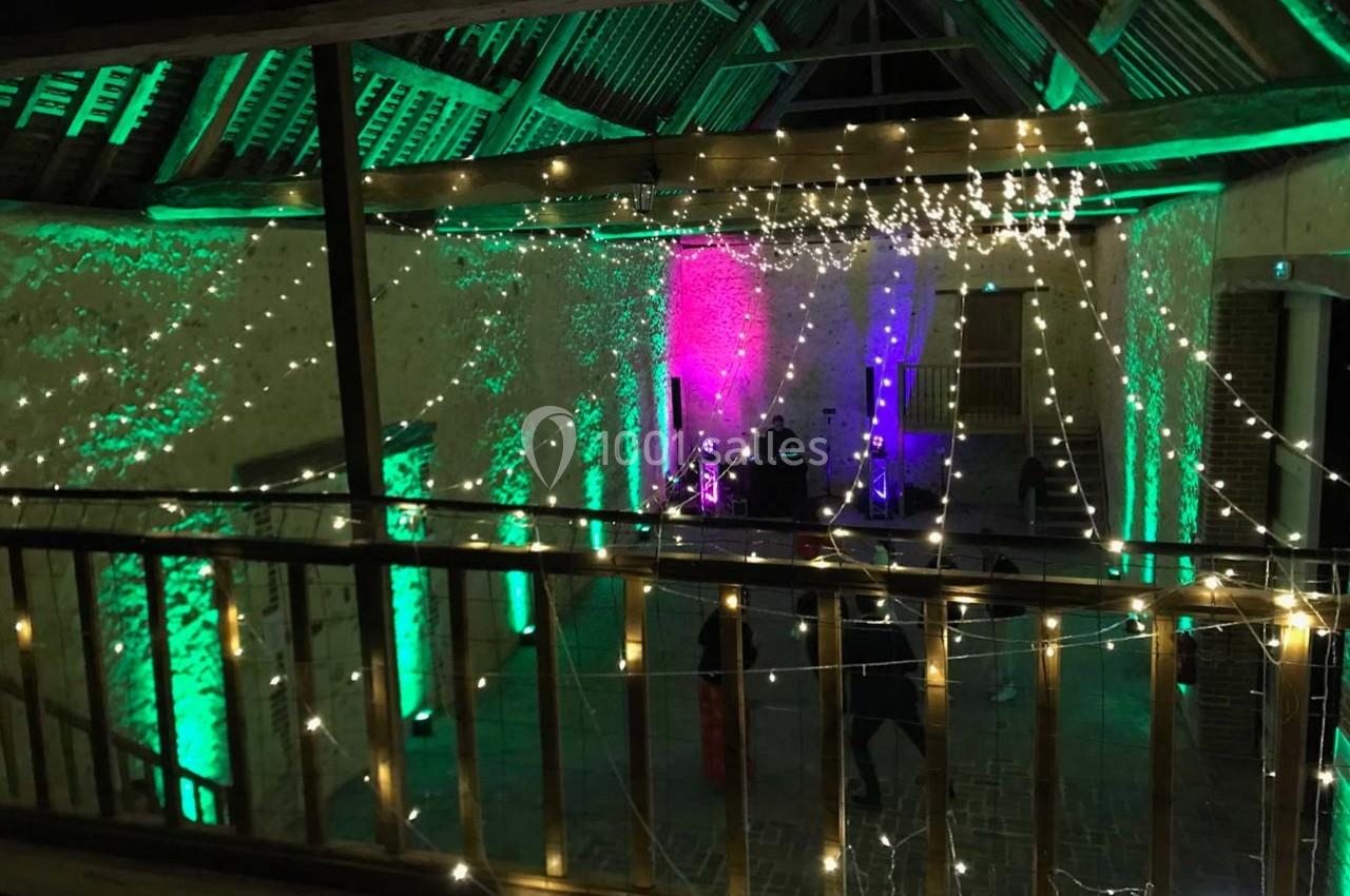 Salle de réception avec charpente en bois, éclairée par des guirlandes lumineuses et des projecteurs colorés.