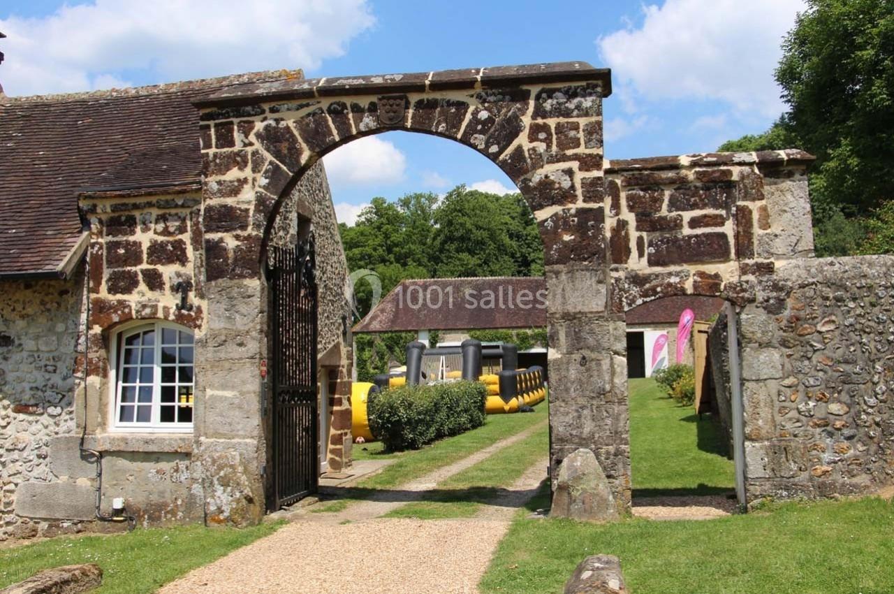 Arc en pierre avec portail métallique, entouré de bâtiments anciens et d'une pelouse sous un ciel ensoleillé.