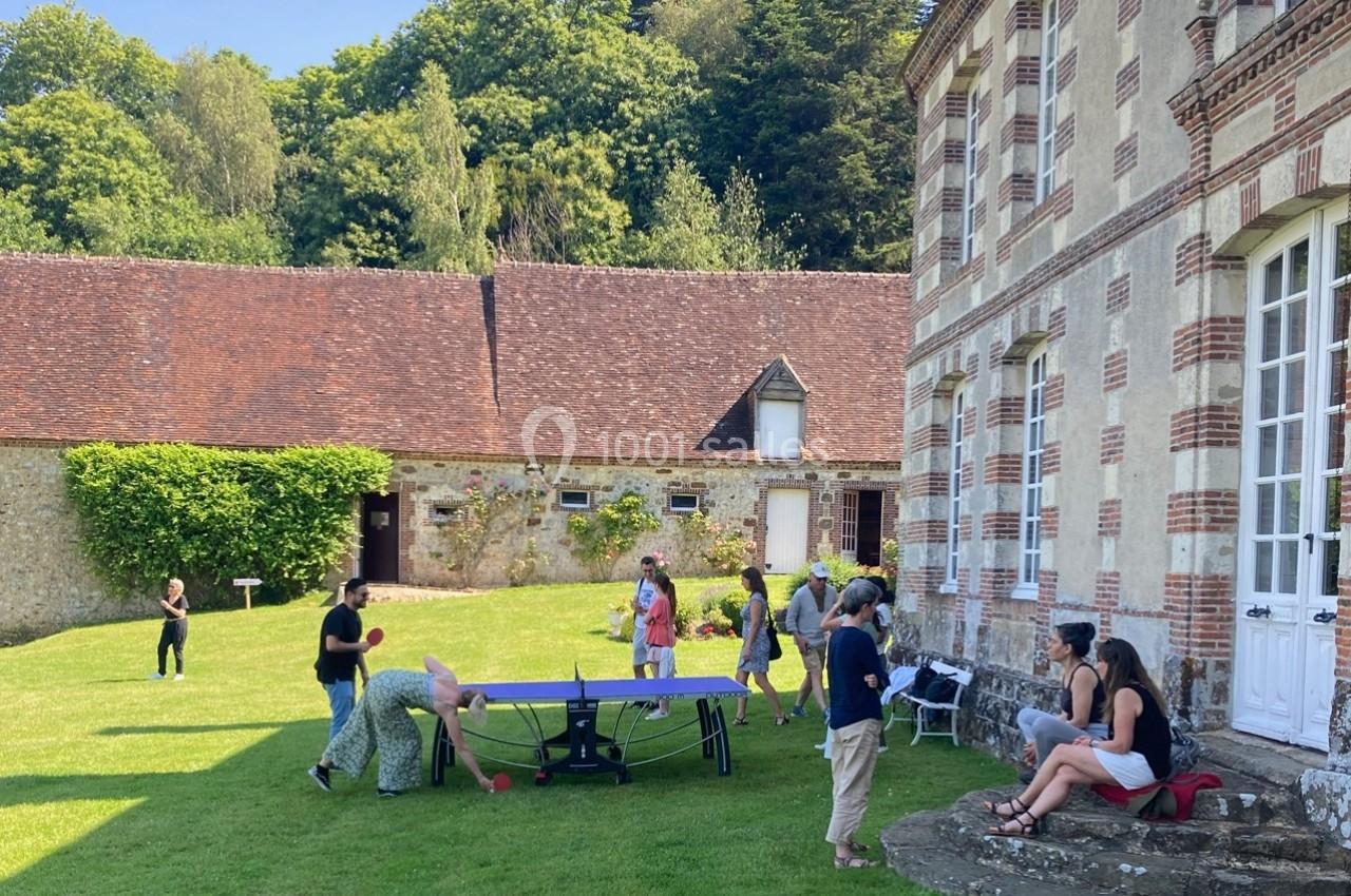 Des personnes jouent au ping-pong et discutent dans le jardin d'une grande maison en pierre, par une journée ensoleillée.