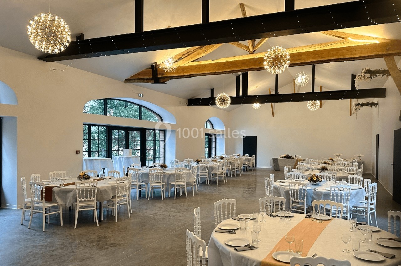Salle de réception lumineuse avec tables dressées, chaises blanches et poutres apparentes sous des luminaires suspendus.