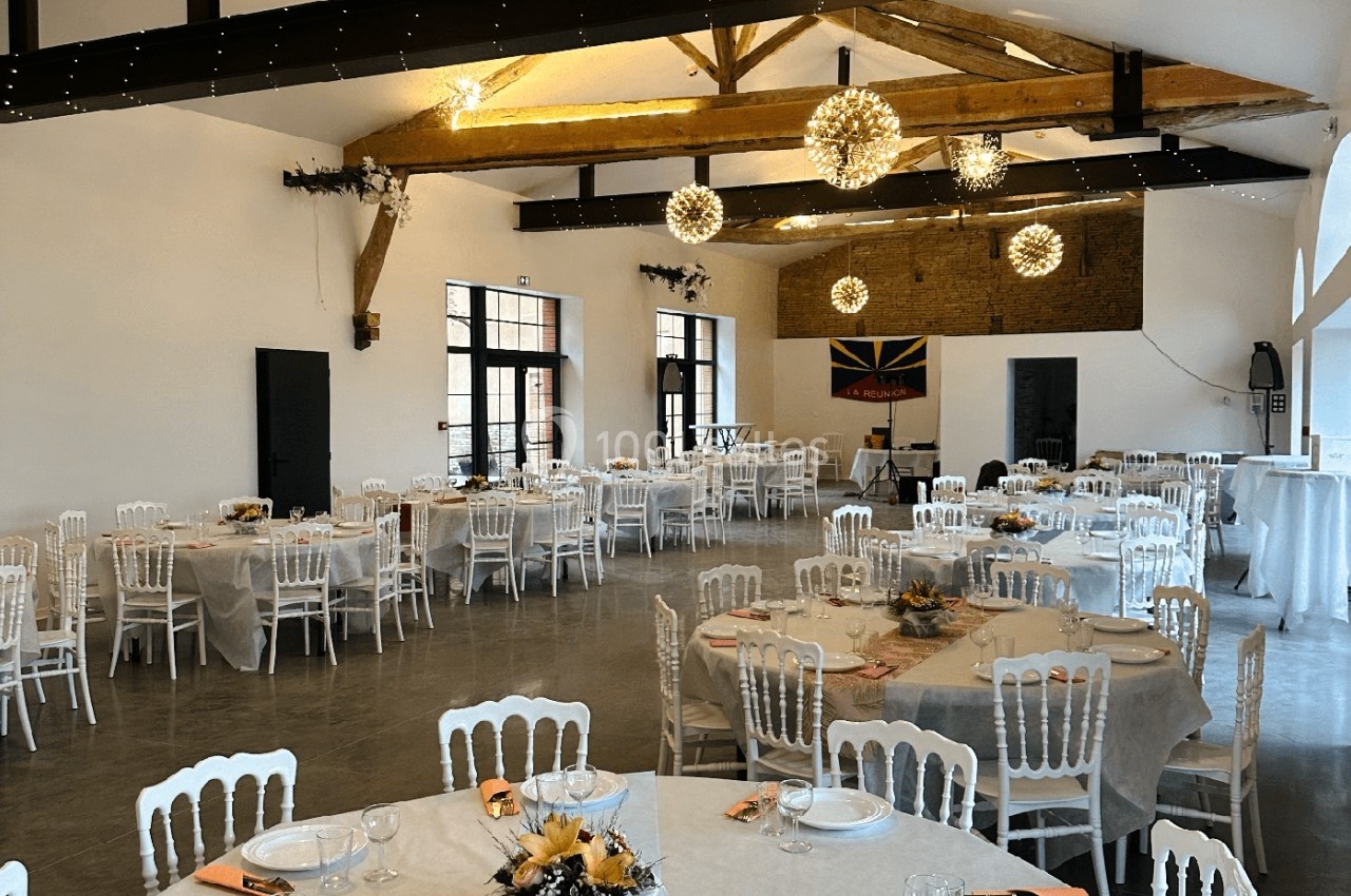 Salle de réception décorée avec des tables rondes dressées, des chaises blanches et des luminaires suspendus.