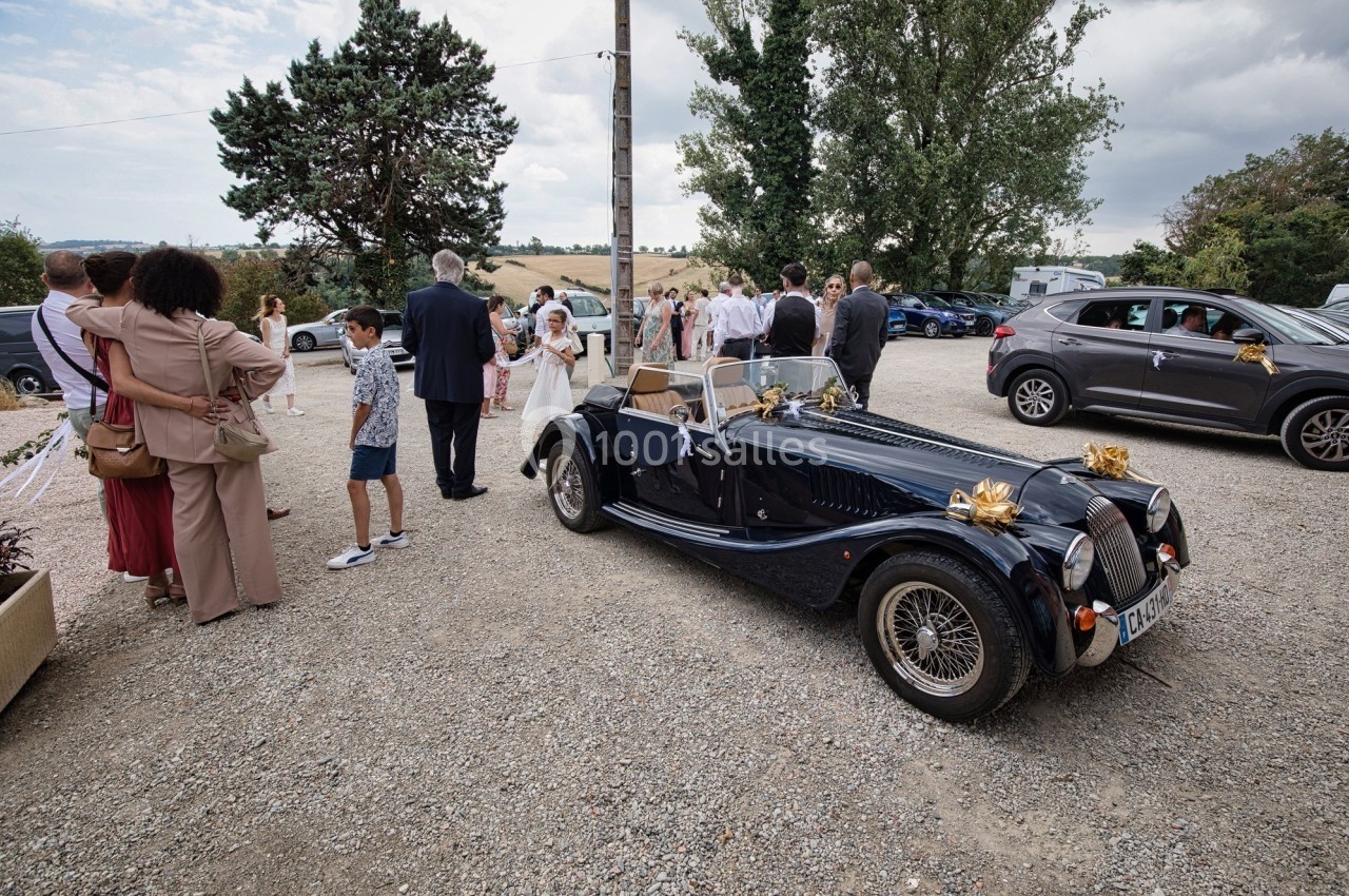 Voiture ancienne décorée pour un mariage, entourée d'invités dans une cour en plein air.