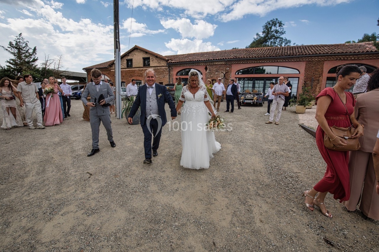 Une mariée en robe blanche marche aux côtés de deux hommes devant un bâtiment en briques, entourée d'invités.