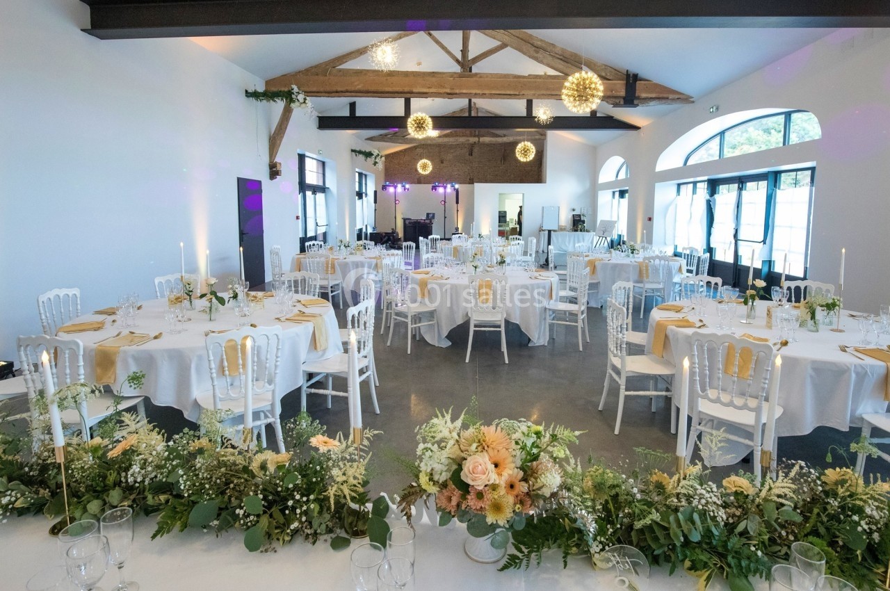 Salle de réception lumineuse décorée avec des tables rondes, nappes blanches, chaises élégantes et arrangements floraux.
