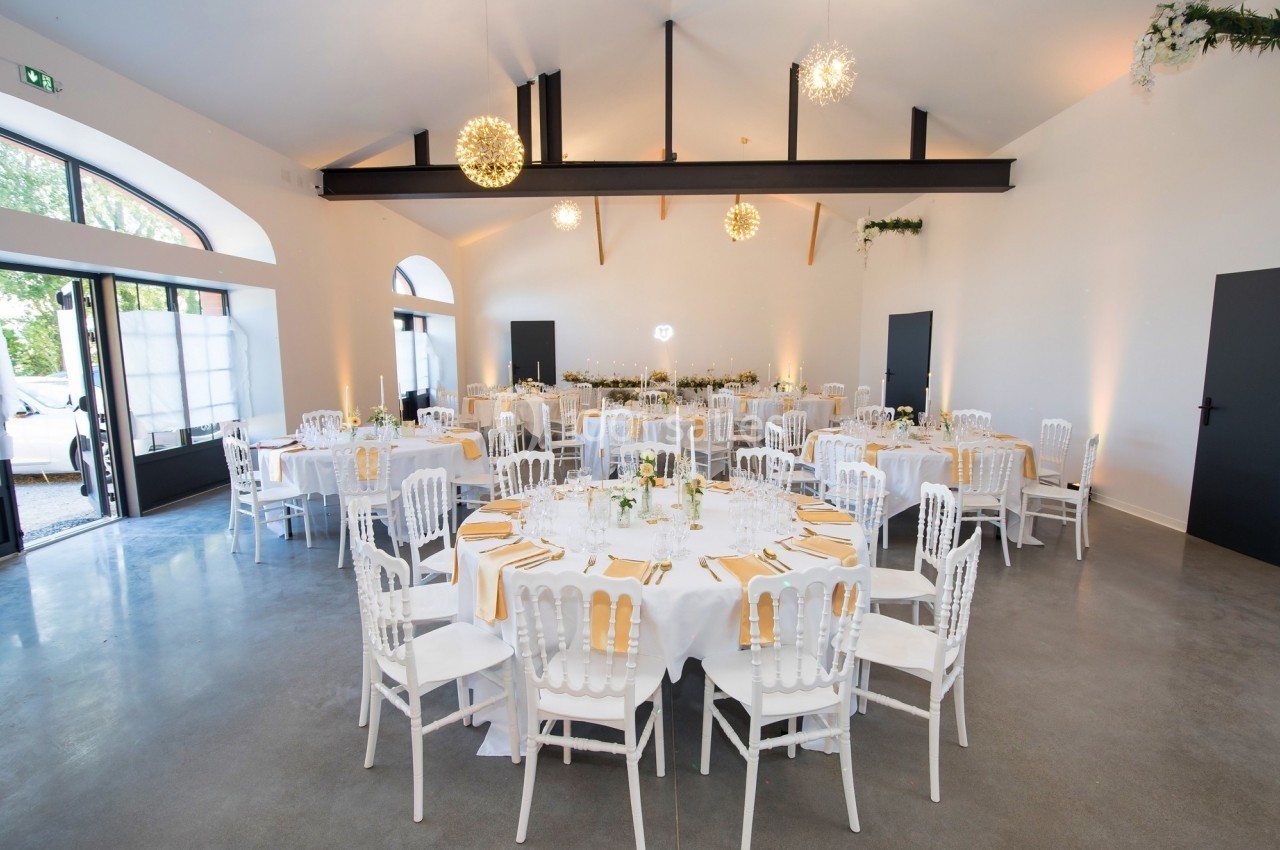 Salle de réception lumineuse avec tables rondes dressées, chaises blanches et décorations élégantes.