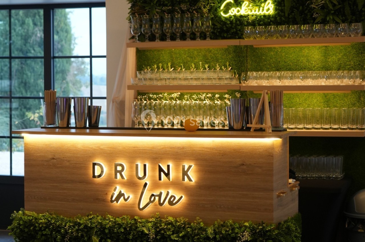 Bar en bois éclairé avec l'inscription ’Drunk in Love’, décoré de verdure et de verres alignés sur des étagères.