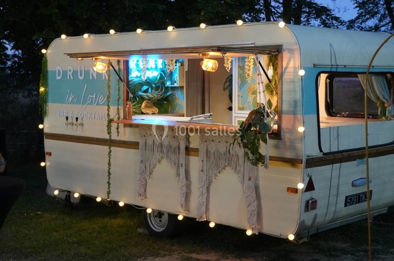 Caravane aménagée en bar lumineux avec guirlandes, plantes suspendues et décorations bohèmes, installée en extérieur.