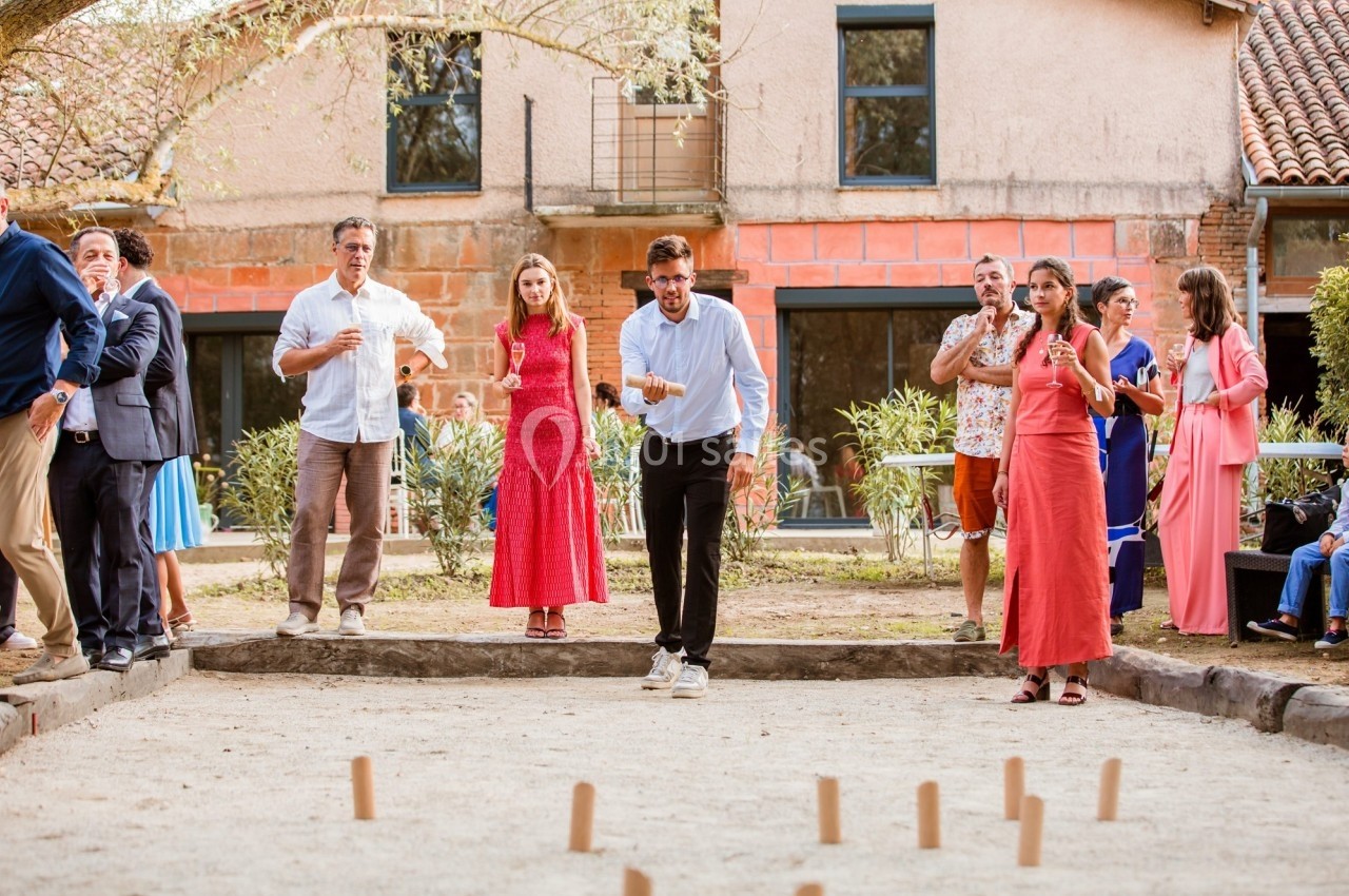 Des personnes jouent à un jeu de quilles en bois dans un jardin, entourées de spectateurs près d'une maison.