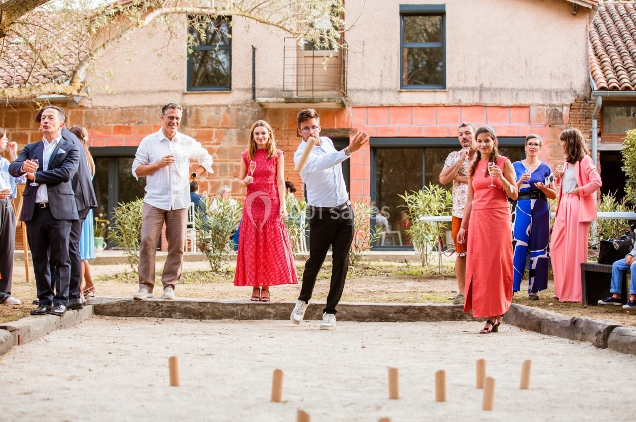 Des personnes jouent à un jeu de quilles en bois en extérieur, entourées de spectateurs devant une maison.
