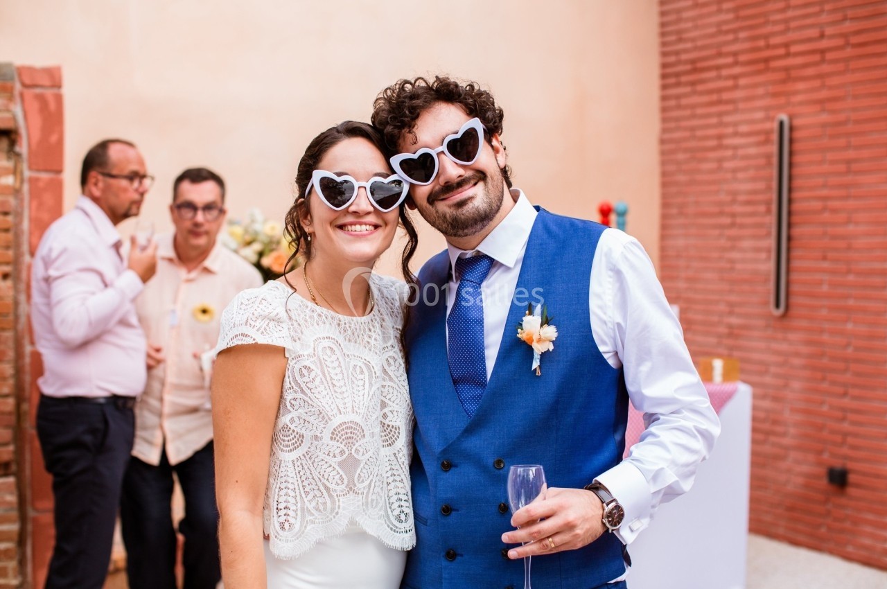 Un couple souriant portant des lunettes en forme de cœur pose lors d'un événement festif en extérieur.