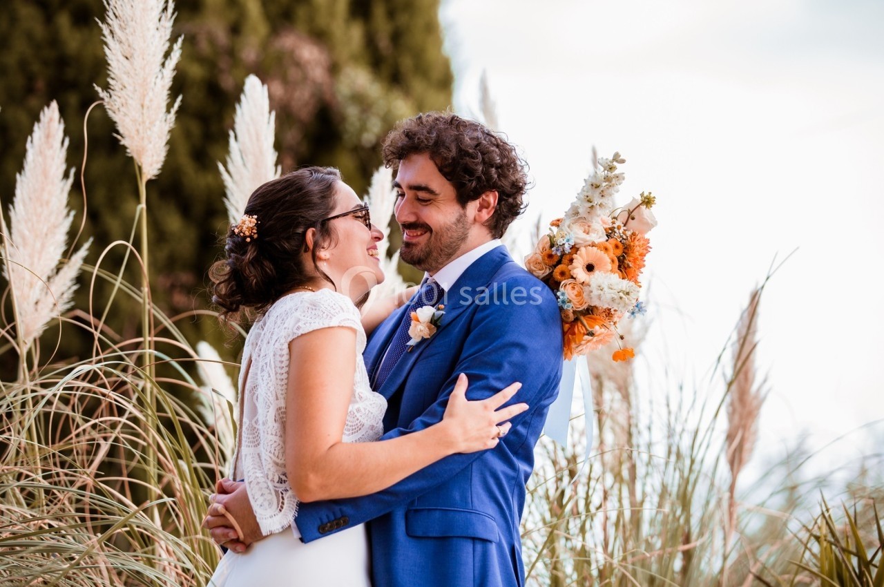 Un couple en tenue de mariage s'enlace dans un jardin avec des herbes hautes et un bouquet de fleurs.